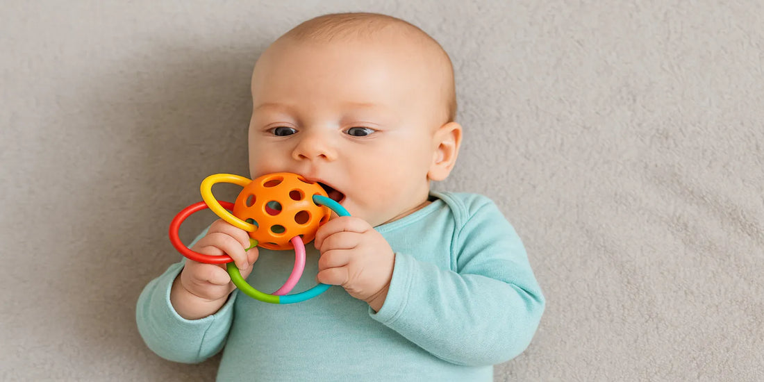 A 3-month-old baby in a turquoise onesie lying on a blanket, holding and chewing a colorful BPA-free sensory teether toy designed for safe play.